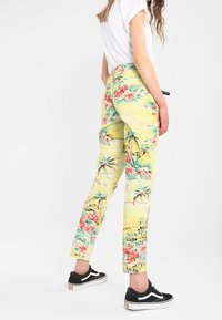 Gula mönstrade byxor med blommönster i rosa och grönt, slim fit, med sidofickor, bärs med en vit t-shirt och svarta sneakers.