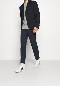 Blazer sombre sur un pull léger à texture, associé à un pantalon slim noir et des baskets blanches avec des accents noirs.