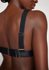 Cinghia di reggiseno nera a coste vista da dietro, con fibbia regolabile in tonalità oro e texture della pelle liscia. Design minimalista.