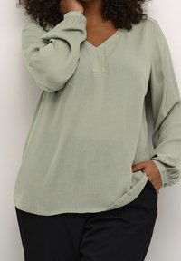 Hellgrüne Bluse mit V-Ausschnitt und langen, puffärmel. Hergestellt aus strukturiertem Stoff, mit einem subtilen Nähedetail und lockerer Passform.