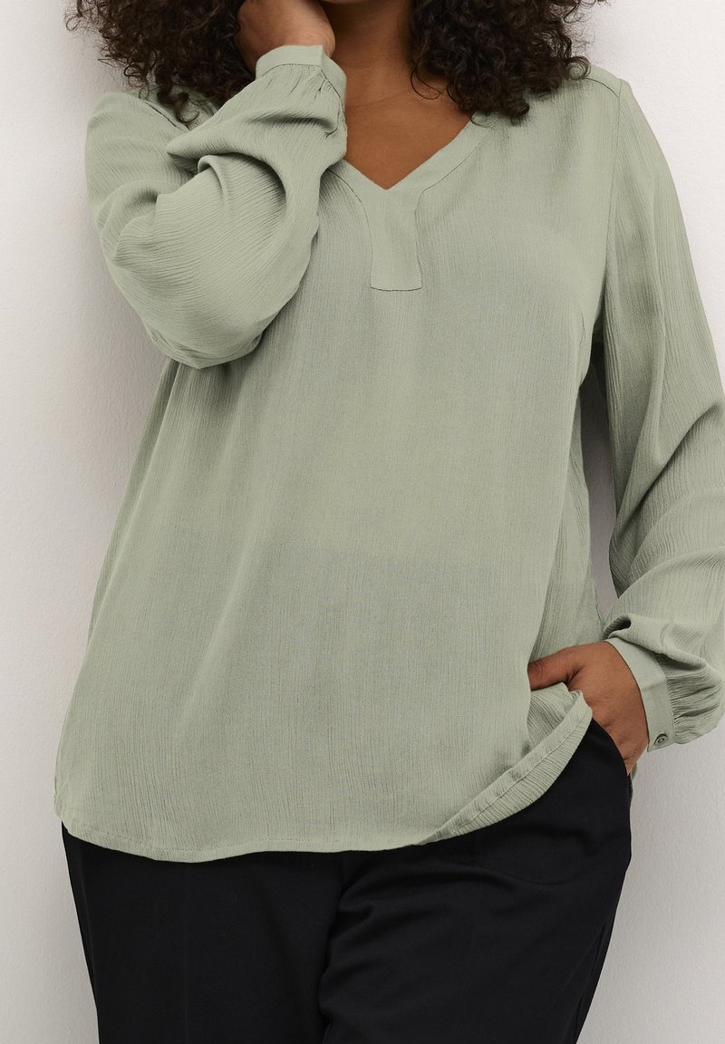 Hellgrüne Bluse mit V-Ausschnitt und langen, puffärmel. Hergestellt aus strukturiertem Stoff, mit einem subtilen Nähedetail und lockerer Passform.