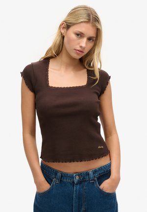 Braunes geripptes Crop-Top mit quadratischem Halsausschnitt, gewellten Kanten und kurzen Ärmeln. Goldenes Logo-Detail am Saum. Kombiniert mit hochgeschnittenen Jeans.