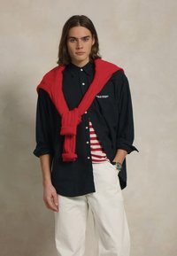 Camisa de botones azul marino oscura con un suéter rojo atado alrededor de los hombros, pantalones blancos y una camiseta a rayas roja y blanca debajo.