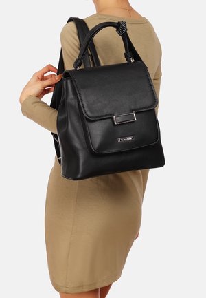Femme en robe beige tenant un sac à dos en cuir noir avec rabat et fermoir métallique sur l'épaule.