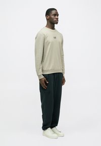 Μπεζ ανοιχτό μακρυμάνικο φούτερ με λογότυπο, συνδυασμένο με σκουροπράσινο φαρδύ παντελόνι και λευκά αθλητικά παπούτσια. Ομαλή υφή, άνετη εφαρμογή.