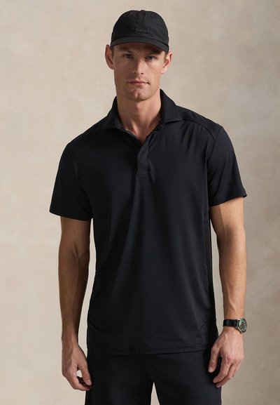 Μαύρο πόλο πουκάμισο με γιακά, κοντά μανίκια και άνετη εφαρμογή· κατασκευασμένο από μαλακό, αεριζόμενο ύφασμα, φορεμένο με ταιριαστό μαύρο καπέλο.