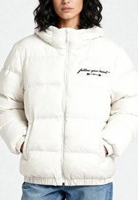 Doudoune blanche avec fermeture éclair à l'avant, design matelassé et capuche. Présente un texte brodé "suis ton cœur" en noir sur la poitrine.