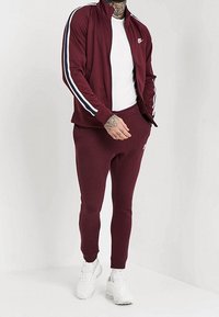 Bordowy dres z białymi i granatowymi paskami, składający się z kurtki na zamek i dopasowanych spodni dresowych, wykonany z teksturowanej tkaniny, zestawiony z białymi sneakersami.