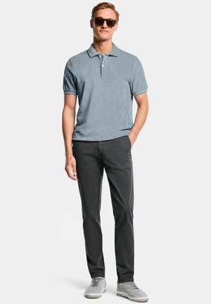 Homme portant des lunettes de soleil, un polo à manches courtes bleu clair, un pantalon gris foncé et des baskets grises, debout avec une main dans la poche.