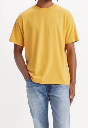 Homme portant un t-shirt Levi's jaune moutarde et un jean bleu clair, debout avec une main dans la poche sur fond blanc.