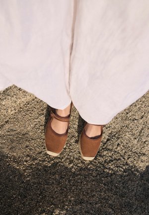 Persona que lleva pantalones de color claro y zapatos planos marrones de punta cerrada, de pie sobre un suelo de grava con textura.