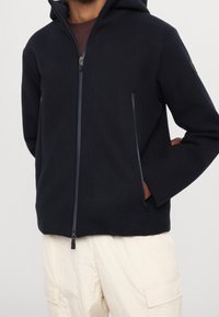 Kapsáč s kapucí v tmavě námořnické barvě, vyrobený z měkké látky. Obsahuje dvě boční kapsy na zip a přední zipové zapínání. Uvolněný střih, minimalistický design.