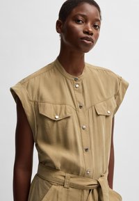 Khaki sleeveless shirt with a buttoned placket, two chest pockets, and a belted waist. Fabric appears lightweight and textured.