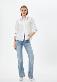 Camicia bianca con bottoni realizzata in tessuto testurizzato, con ricami blu floreali sul colletto e sui polsini, abbinata a jeans chiari svasati.