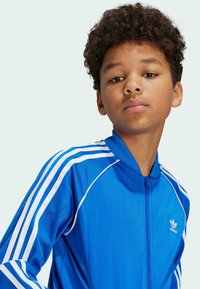 Modrá bunda na zips vyrobená z hladkého materiálu, s bielymi pruhovanými akcentmi na rukávoch a malým bielym logom Adidas na hrudi.