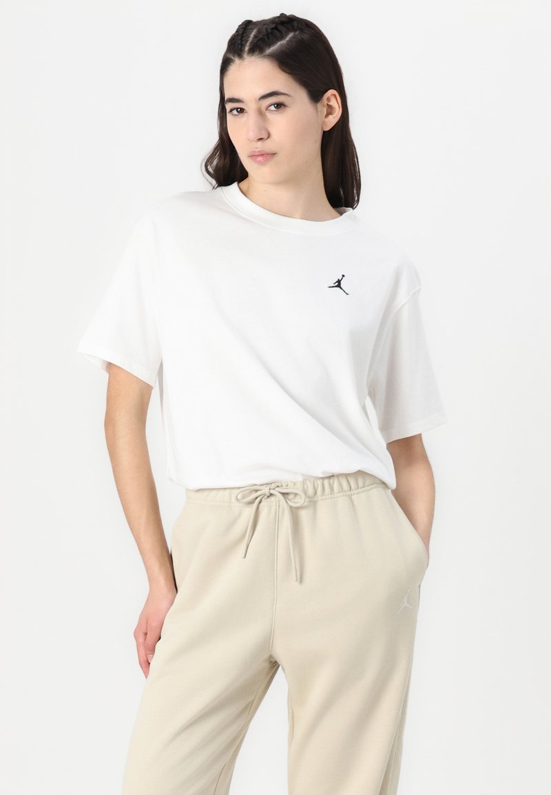 Weißes Baumwoll-T-Shirt mit rundem Halsausschnitt und kurzen Ärmeln, das ein kleines schwarzes Logo auf der Brust trägt. Kombiniert mit beigen Jogginghosen mit Kordelzug.