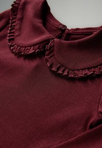 Burgundy ribbat tyg topp med rundad krage som har volangkanter och en knappstängning bak.