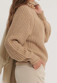 Světle béžový pletený svetr s texturovaným žebrovaným vzorem a volnými rukávy, doplněný krémovými kalhotami. Výrazný řetízkový detail na výstřihu.