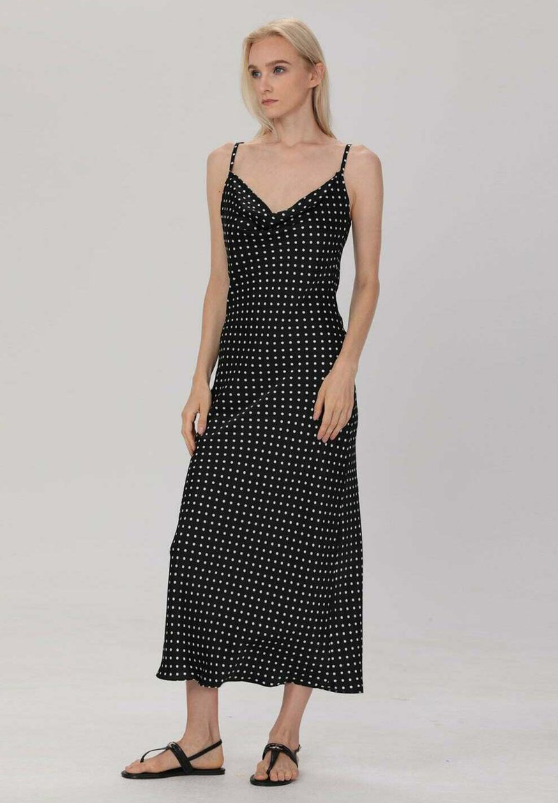 Robe maxi noire à pois blancs, avec des bretelles fines et un décolleté en cowl. Le tissu a une texture lisse et une silhouette fluide.
