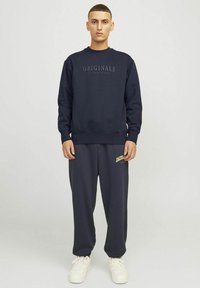 Marine sweatshirt met de tekst "ORIGINALS", gecombineerd met donkergraat broeken met "JACK & JONES" in het geel. Model draagt witte sneakers.