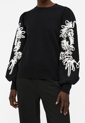 Femme portant un pull noir avec de longues manches bouffantes ornées d'une broderie florale blanche le long de chaque bras.