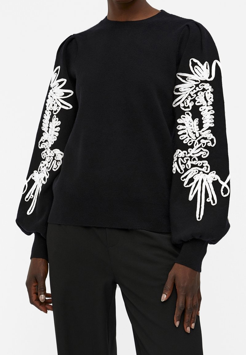 Femme portant un pull noir avec de longues manches bouffantes ornées d'une broderie florale blanche le long de chaque bras.