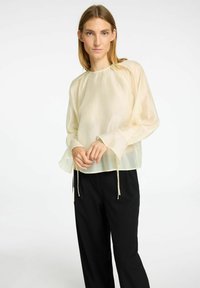 Blusa trasparente di un giallo chiaro con maniche lunghe e ampie e dettagli con cordoncino sul collo, abbinata a pantaloni neri.