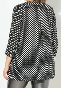Svart tunika med litet vitt hjärtmönster, långa ärmar, rundad nederkant och en plissédetalj på ryggen. Slät textur.