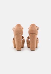 Beige mokkanahkaiset korkokengät, joissa on lohkokorko, nilkkaremmi soljella ja avoin varvasosa. Sileä pinta mattapintaisella viimeistelyllä.