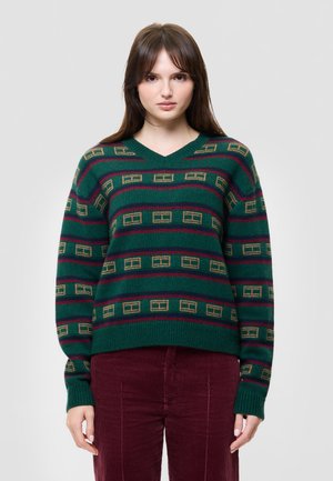 Une femme porte un pull vert à motifs et à col en V, un pantalon évasé bordeaux et des bottines noires devant un fond de studio gris uni.