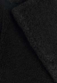 Czarna tkanina polarowa o miękkiej, teksturowanej powierzchni. Charakteryzuje się grubą, kręconą fakturą z gładkim brzegiem, odpowiednią do odzieży wierzchniej.