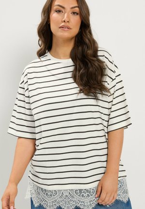 Kobieta z długimi falistymi włosami, ubrana w biały shirt z krótkim rękawem z czarnymi poziomymi paskami i białą koronkową lamówką na dole.