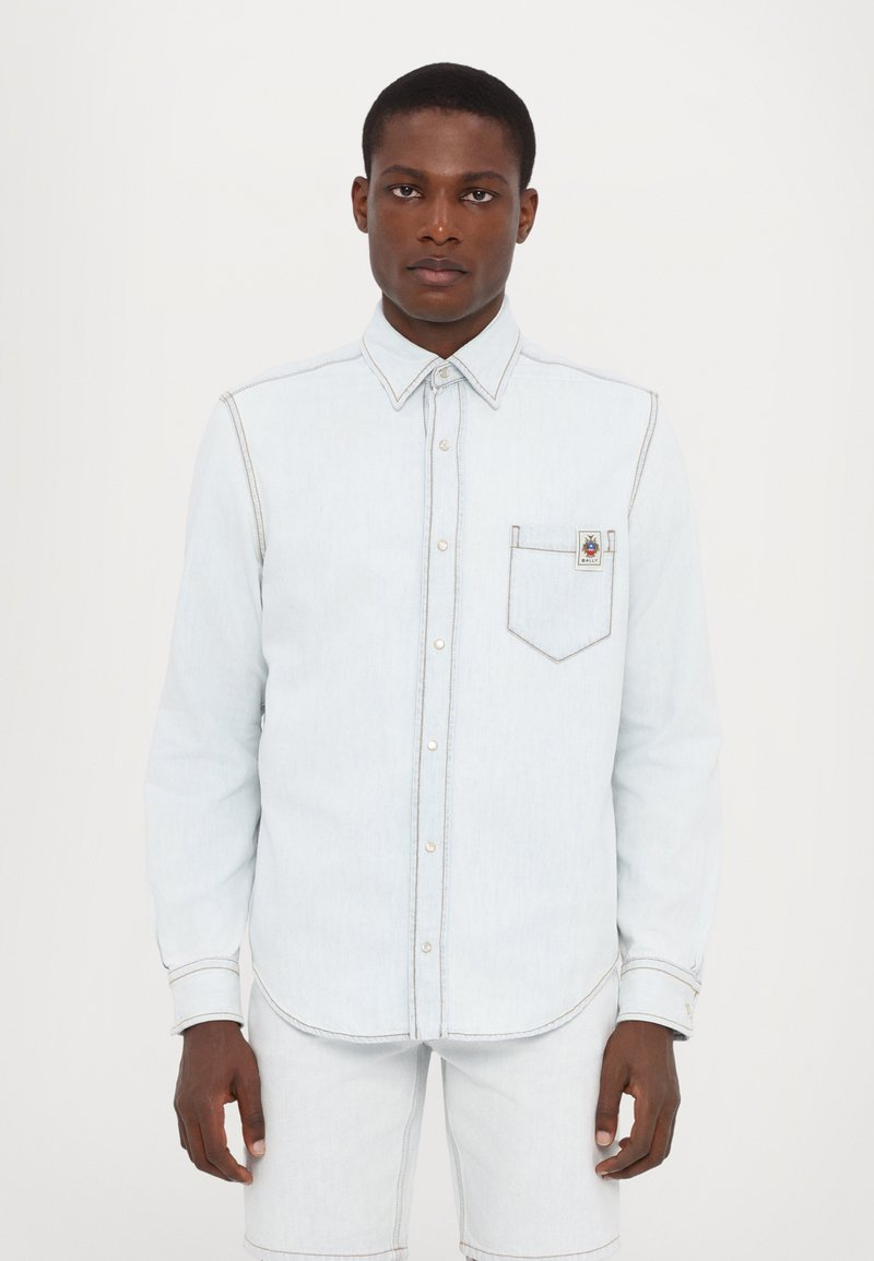 Camicia di jeans azzurro chiaro con maniche lunghe, colletto con bottoni, taschino sul petto, dettagli con cuciture bianche e un'etichetta con il logo sulla tasca.
