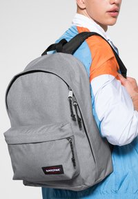 Harmaa Eastpak-reppu, jossa on pääosasto, etupussin vetoketju, mustat vetoketjun vedinsangot ja pehmustetut olkahihnat, jota henkilö käyttää.
