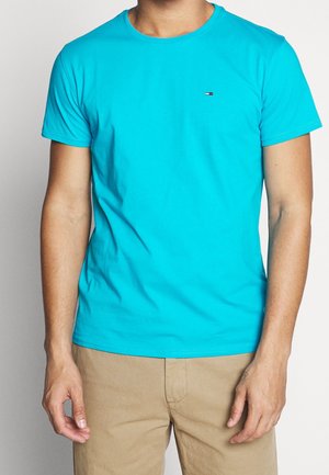 Mężczyzna w jasnoniebieskim krótkim rękawie z małym logo na piersi i beżowych spodniach, stojący na jednolitym tle.