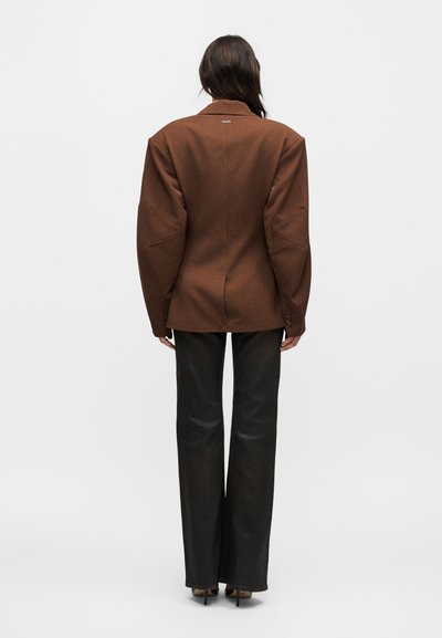 Άτομο με μακριά σκουρόχρωμα μαλλιά φορά υπερμεγέθη καφέ σακάκι και μαύρο παντελόνι με φαρδιά μπατζάκια, στέκεται αντικρίζοντας ένα απλό λευκό φόντο.