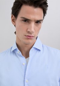 Blau-weiß gestreiftes Hemd mit Knopfleiste und klassischem Kragen. Verfügt über eine glatte Textur und einen dezenten Glanz. Wird über einem hellen T-Shirt getragen.