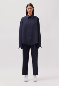 Camicia oversize blu navy con bottoni, colletto aperto e maniche lunghe abbinate a pantaloni coordinati e sneakers bianche.