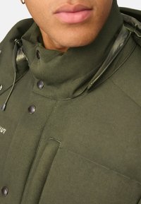Olivengrøn jakke med høj krave, matte sorte trykknapper og en lynlåslomme. Stoffet virker struktureret og robust.