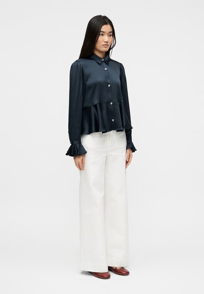 Γυναίκα που στέκεται φορώντας μπλούζα από μπλε σατέν με βολάν στα μανίκια και λευκό παντελόνι με φαρδιά πόδια, συνδυασμένο με καφέ παπούτσια, μπροστά σε απλό φόντο.