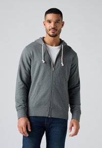 Felpa grigia con zip, dotata di coulisse, tasche frontali e polsini a coste. Indossata sopra una t-shirt bianca, abbinata a jeans blu.