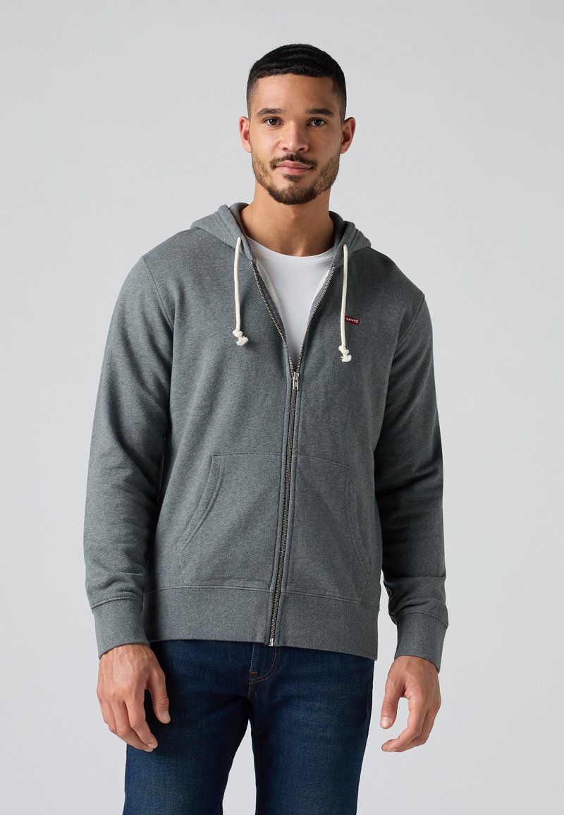 Felpa grigia con zip, dotata di coulisse, tasche frontali e polsini a coste. Indossata sopra una t-shirt bianca, abbinata a jeans blu.