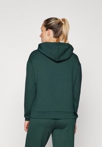 Hoodie vert foncé en tissu doux, doté d'une grande capuche, d'une coupe décontractée et de poignets côtelés. Design simple sans motifs ni accents visibles.