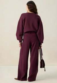 Vrouw met golvend haar draagt een bijpassende diepe paarse losse trui en wijde broek, terwijl ze een kleine paarse handtas aan haar zijde houdt.