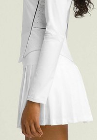 Haut blanc à manches longues avec des panneaux perforés et une coupe ajustée. Associé à une jupe plissée blanche, créant une tenue de sport coordonnée.