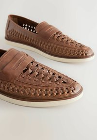 Zapatos de cuero marrón tipo mocasín con un diseño superior tejido, que presentan una textura suave y una suela blanca en contraste. Los acentos incluyen bandas horizontales.