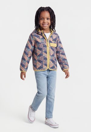 Šypsanti vaikas su pintomis kasomis, dėvintis raštuotą striukę su užtrauktuku, šviesiai mėlynas džinsas ir baltus sportbačius, stovintis su viena koja į priekį.