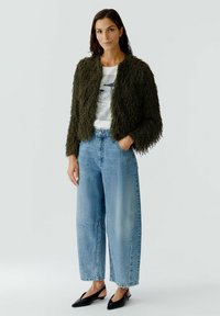 Olive green, fluffy cropped jacket over a white graphic t-shirt. Paired with light blue, wide-leg jeans and black slingback shoes.