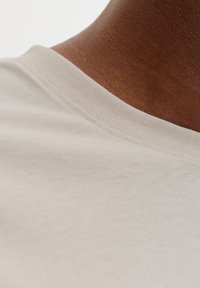 Μπεζ βαμβακερό μπλουζάκι σε ανοιχτή απόχρωση, παρουσιάζοντας κοντινή λήψη στον στρογγυλό λαιμό και την υφή, με ορατές ραφές και απαλά υφάσματα στις λεπτομέρειες του γιακά.