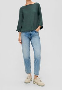 Top de manga larga verde oscuro con textura, combinado con vaqueros de pierna recta azul claro. Zapatillas crema con detalles en verde completan el conjunto.